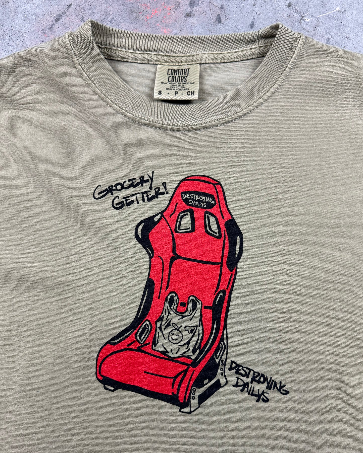 Close up shot of tan 100% cotton t-shirt featuring a hand-drawn red racing seat with a grocery bag and the text "Grocery Getter! Destroying Dailys," perfect for car enthusiasts. Funny car shirt for car enthusiasts