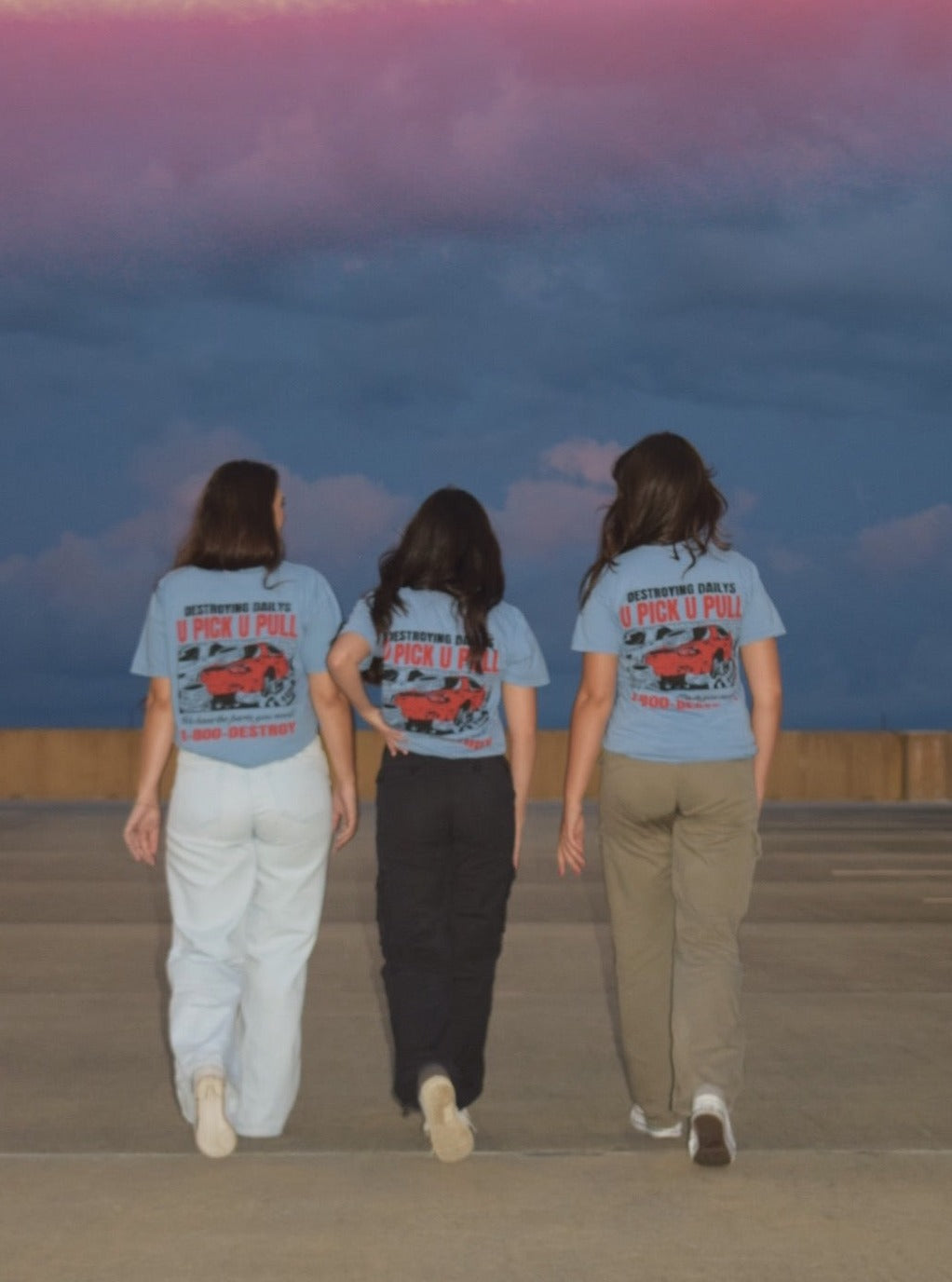 Three women wearing light blue 100% cotton t-shirts with a hand-drawn JDM Rx-7 graphic and text "Destroying Dailys U Pick U Pull 1-800-DESTROY" on the back, ideal for car enthusiasts.