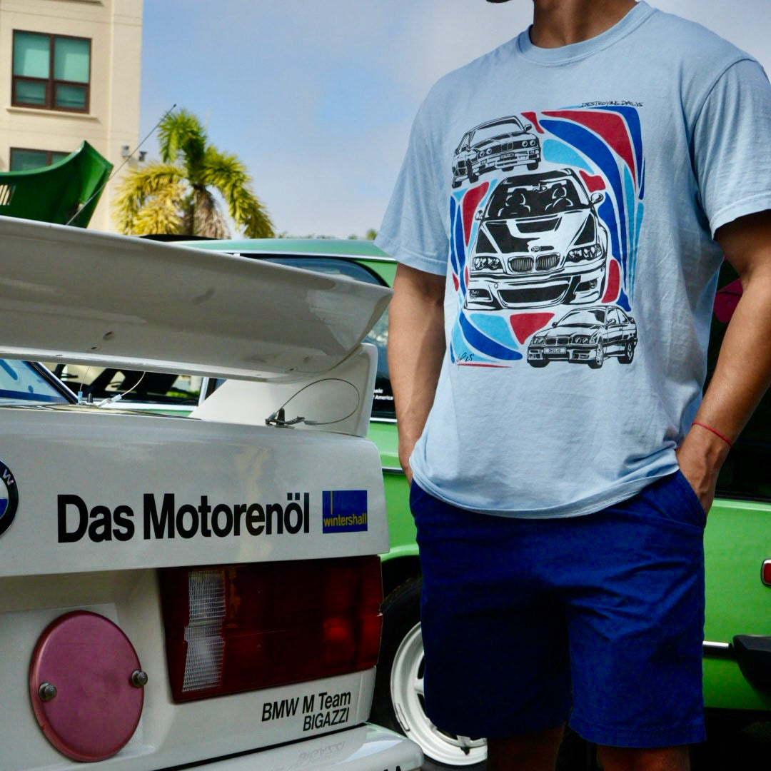 Man wearing a light blue 100% cotton t-shirt featuring hand-drawn BMW E30, E36, and E46 designs with red, blue, and black accents, perfect for car enthusiasts.