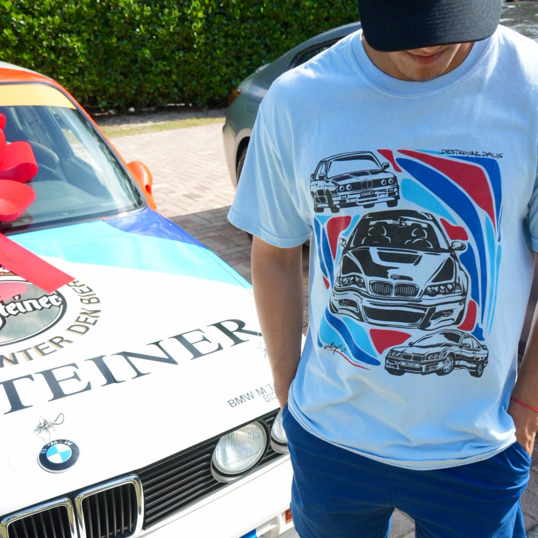 Man wearing a light blue 100% cotton t-shirt featuring hand-drawn BMW E30, E36, and E46 designs with red, blue, and black accents, perfect for car enthusiasts. With Bmw E30 Racecar in the back