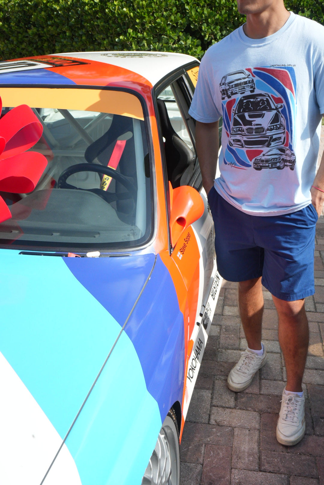 Man wearing a light blue 100% cotton t-shirt featuring hand-drawn BMW E30, E36, and E46 designs with red, blue, and black accents, perfect for car enthusiasts.