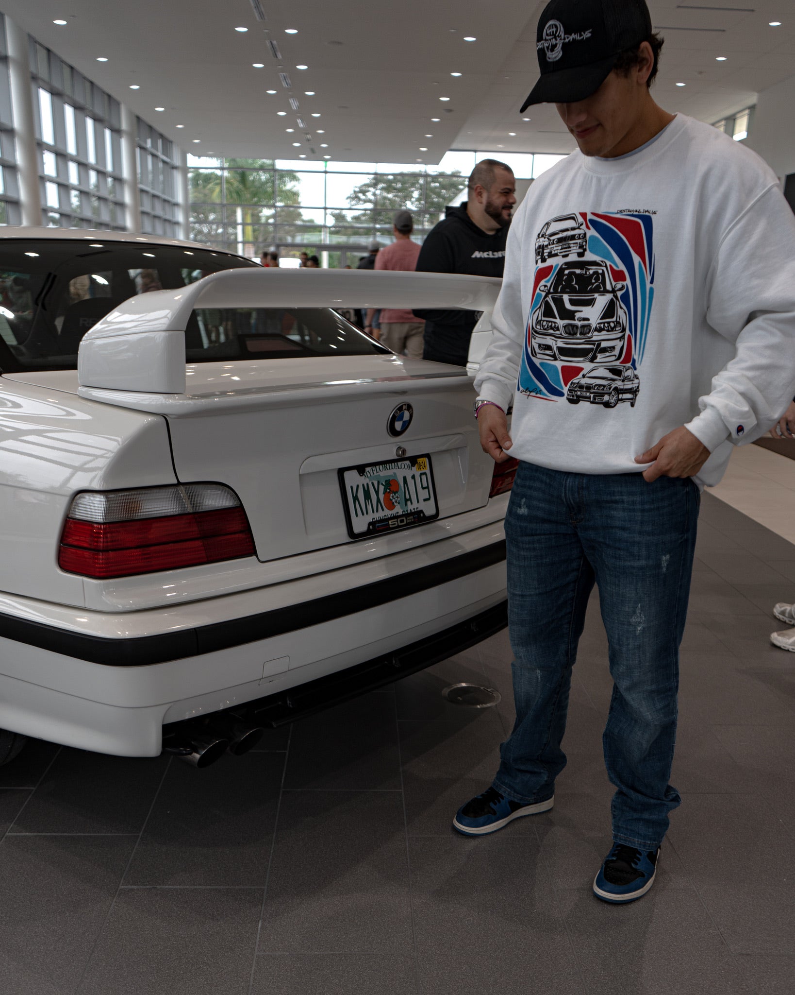 male model at a car meet with a bmw m3 wearing White crewneck sweatshirt made of 50/50 cotton and polyester blend featuring hand-drawn BMW E30, E36, and E46 M3s with red, blue, and black accents, perfect for car enthusiasts. Bmw sweatshirt for car guys