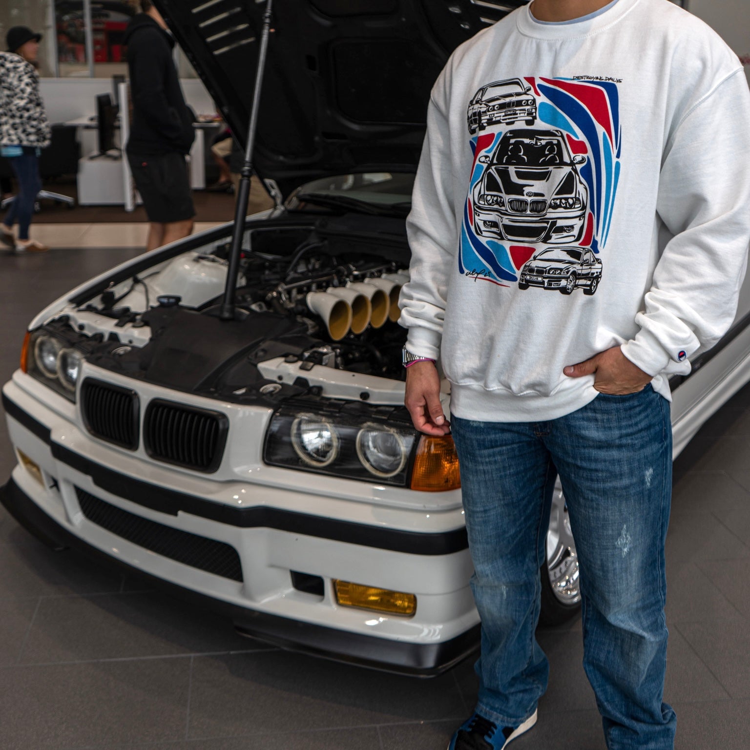 Male model with a BMW m3 wearing White crewneck sweatshirt made of 50/50 cotton and polyester blend featuring hand-drawn BMW E30, E36, and E46 M3s with red, blue, and black accents, perfect for car enthusiasts. Bmw sweatshirt for car guys