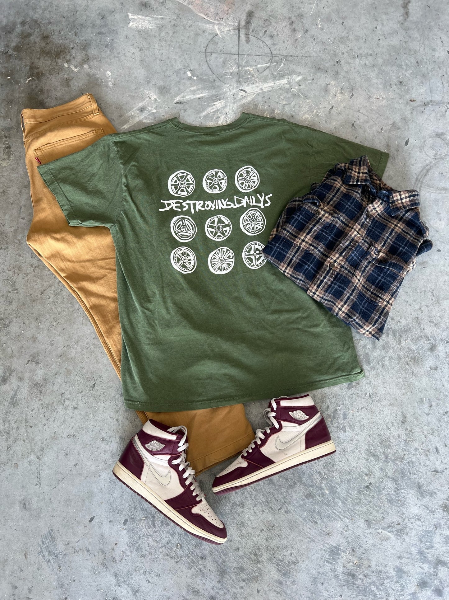 Green 100% cotton t-shirt featuring a hand-drawn JDM wheel design with the text "Destroying Dailys," styled with tan pants, plaid shirt, and maroon sneakers, perfect for car enthusiasts.