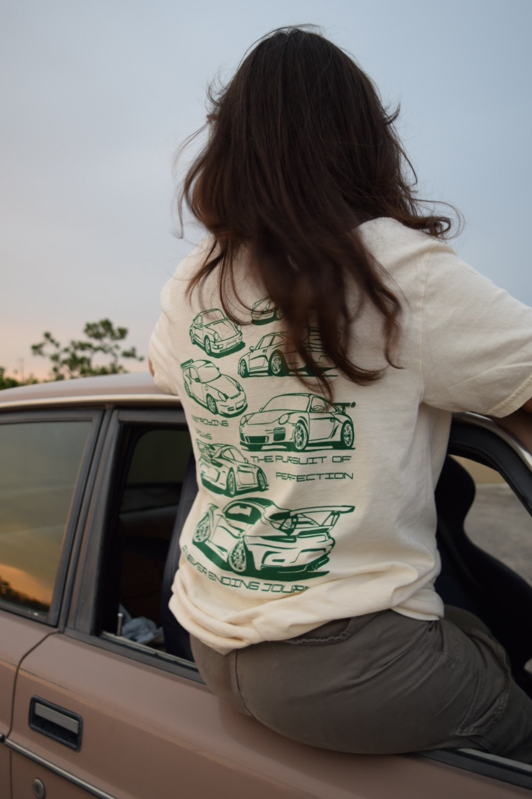 Female Model In car wearing White 100% cotton t-shirt featuring green hand-drawn Porsche 911 GT3 RS designs with the text "Destroying Dailys The Pursuit of Perfection A Never Ending Journey," perfect for car enthusiasts. Porsche Tee for porsche guys