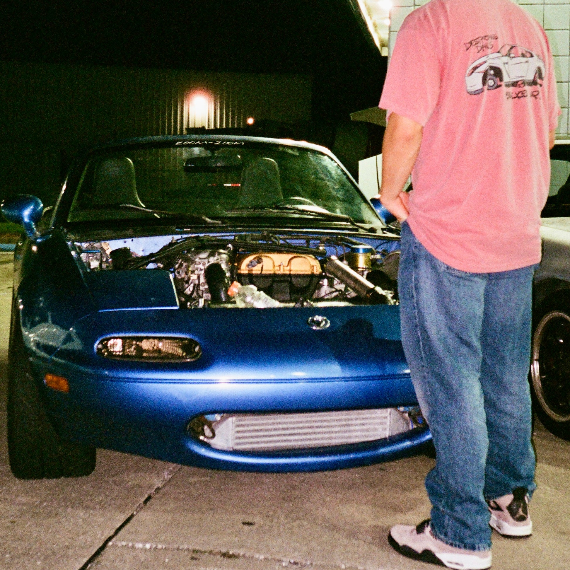 model wearing Red 100% cotton t-shirt featuring a hand-drawn Nissan GTR on bricks with the text "Destroying Dailys Bricked Up!" perfect for JDM enthusiasts. Funny Tee for Car guys, in front of miata