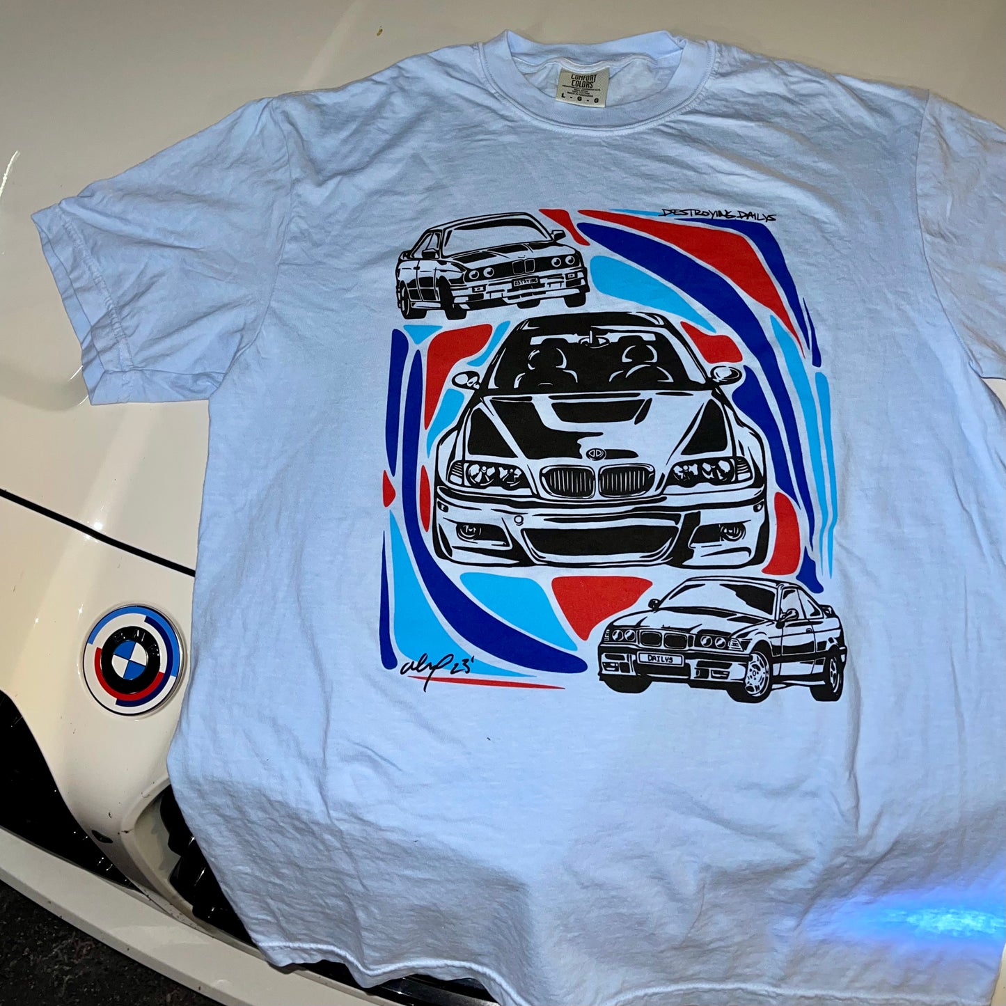 Light blue 100% cotton t-shirt featuring hand-drawn BMW E30, E36, and E46 designs with red, blue, and black accents, ideal for car enthusiasts. on bmw m3 lifestyle photo