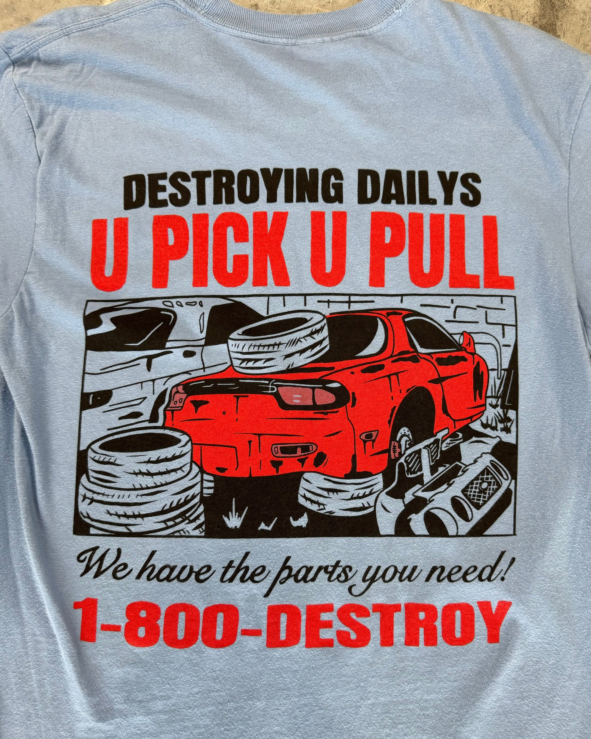 Light blue 100% cotton t-shirt featuring a hand-drawn JDM Rx-7 at a scrapyard with the text "Destroying Dailys U Pick U Pull 1-800-DESTROY," ideal for car enthusiasts, Close up detailed photo of the hand drawn JDM RX-7 Design