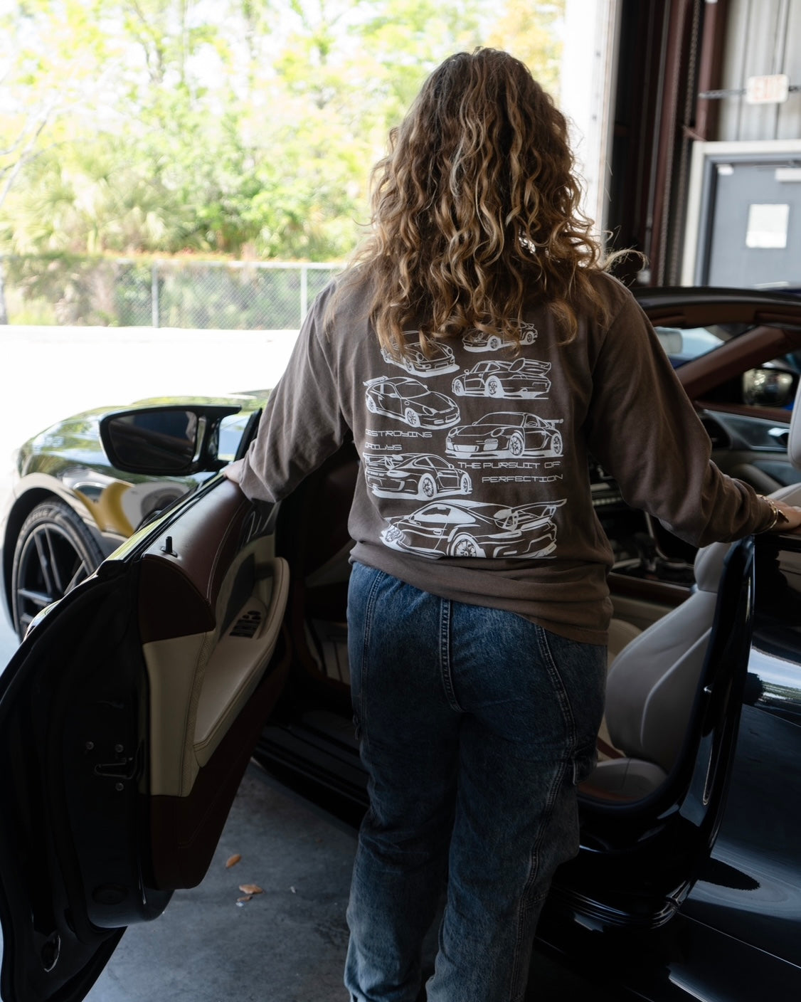 Female model wearing Brown long-sleeve 100% cotton t-shirt featuring white hand-drawn Porsche 911 GT3 RS designs with the text "Destroying Dailys The Pursuit of Perfection A Never Ending Journey," perfect for car enthusiasts.