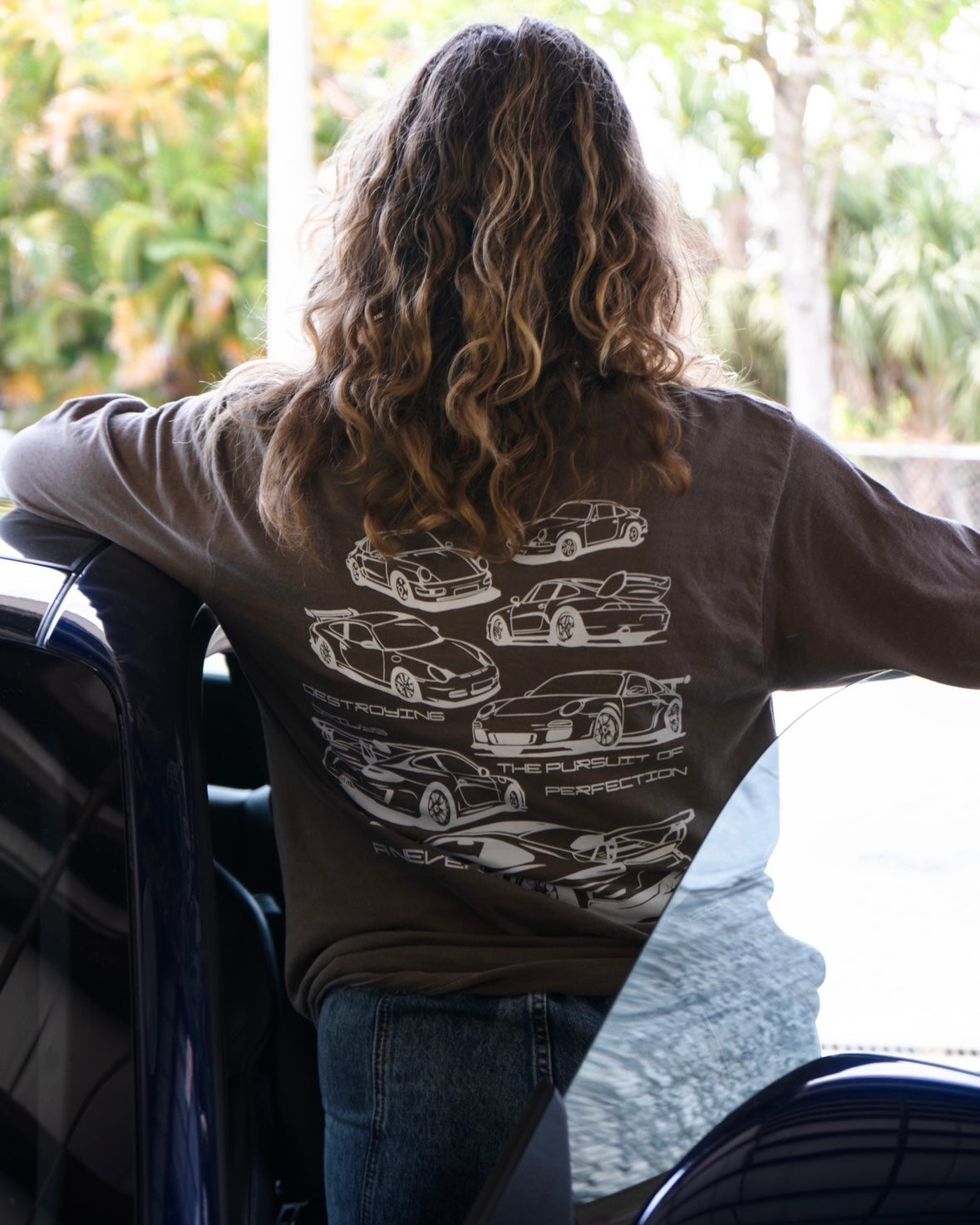 Female model wearing Brown long-sleeve 100% cotton t-shirt featuring white hand-drawn Porsche 911 GT3 RS designs with the text "Destroying Dailys The Pursuit of Perfection A Never Ending Journey," perfect for car enthusiasts.