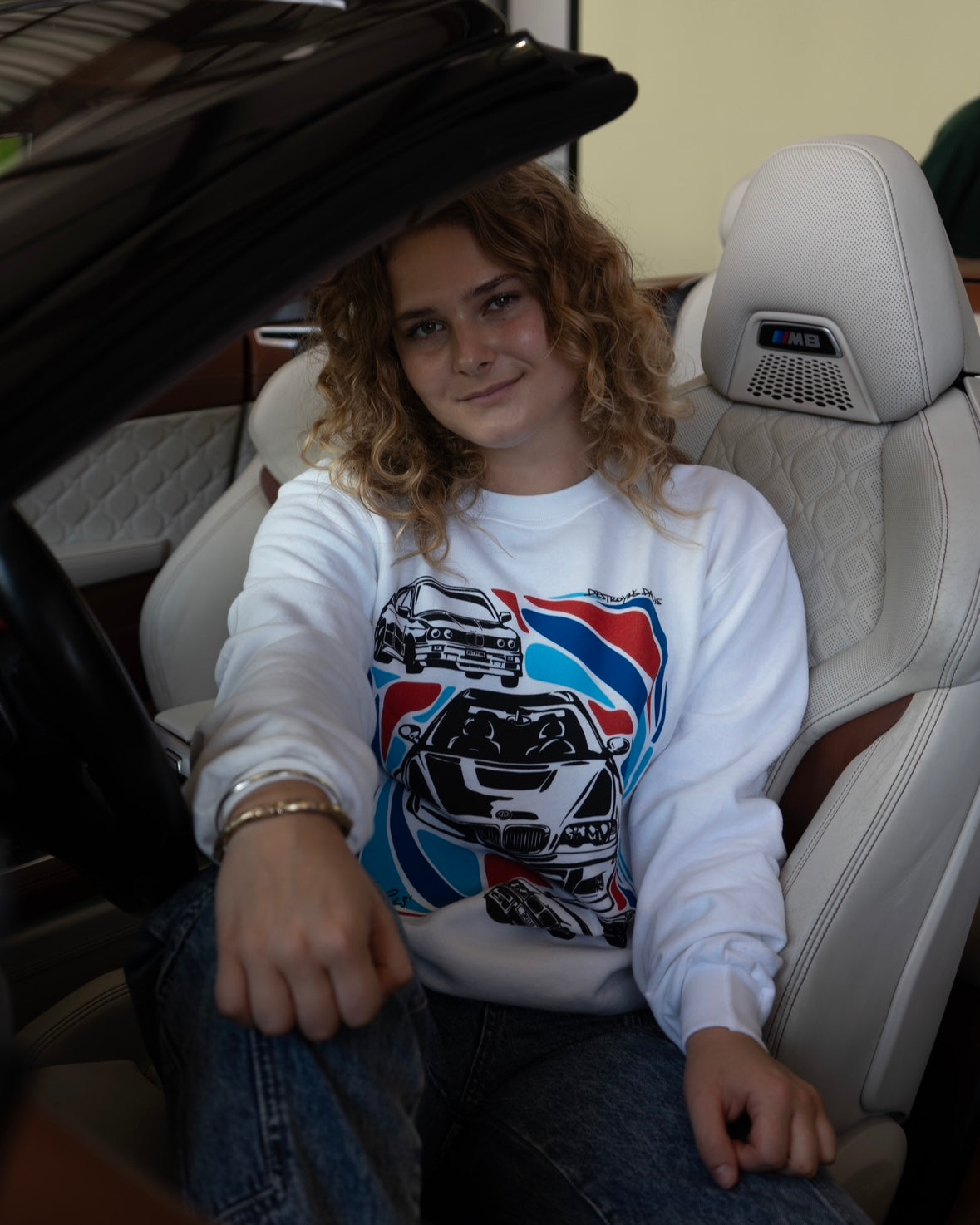 female model in a BMW M8 wearing a White crewneck sweatshirt made of 50/50 cotton and polyester blend featuring hand-drawn BMW E30, E36, and E46 M3s with red, blue, and black accents, perfect for car enthusiasts. Bmw sweatshirt for car guys