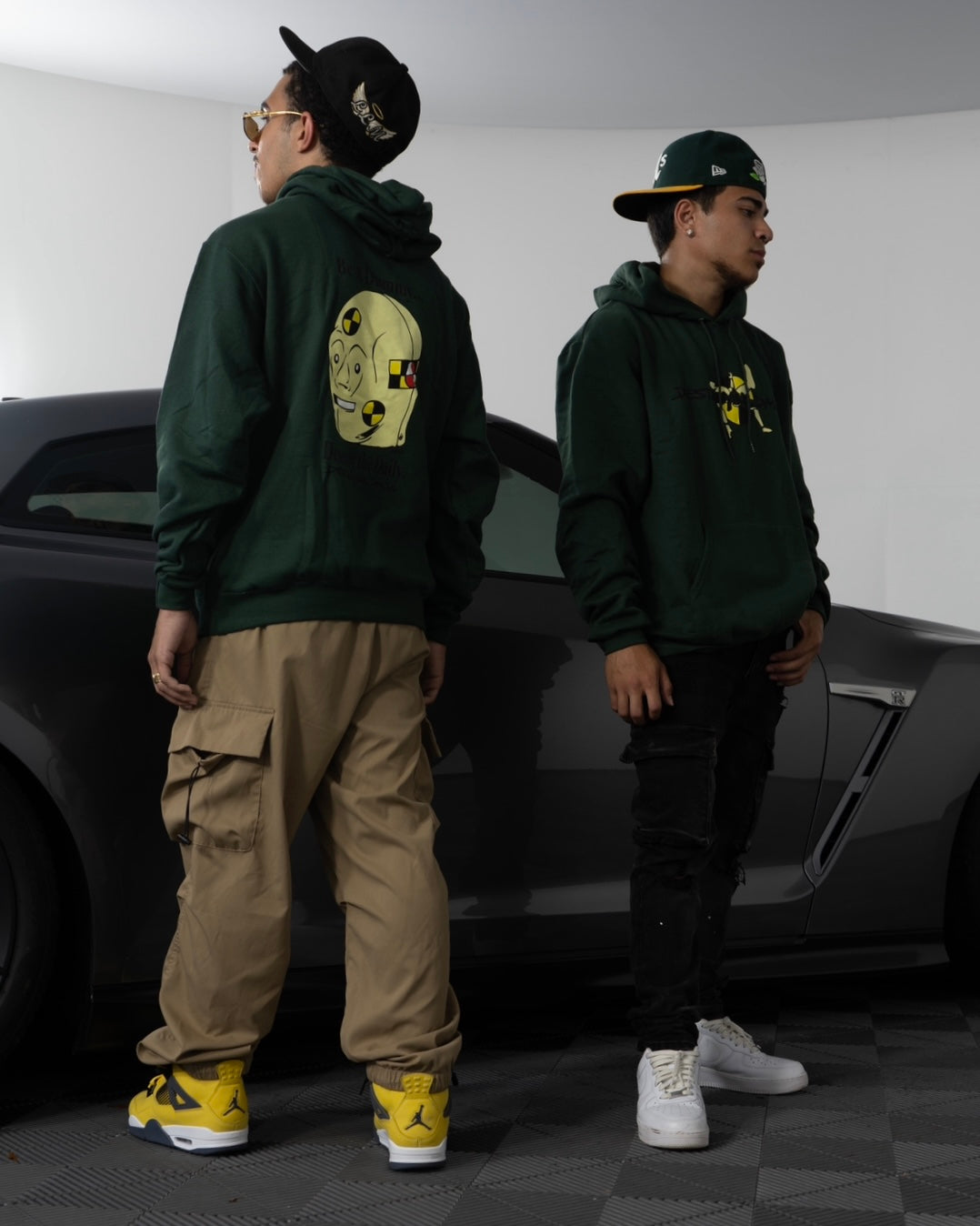 Male streetwear models wearing Dark green hoodie made of 50/50 cotton and polyester blend featuring a hand-drawn crash test dummy with the text "Be A Dummy... Destroy the Daily: Destroying Dailys," perfect for modified car enthusiasts. funny hoodie for car enthusiasts