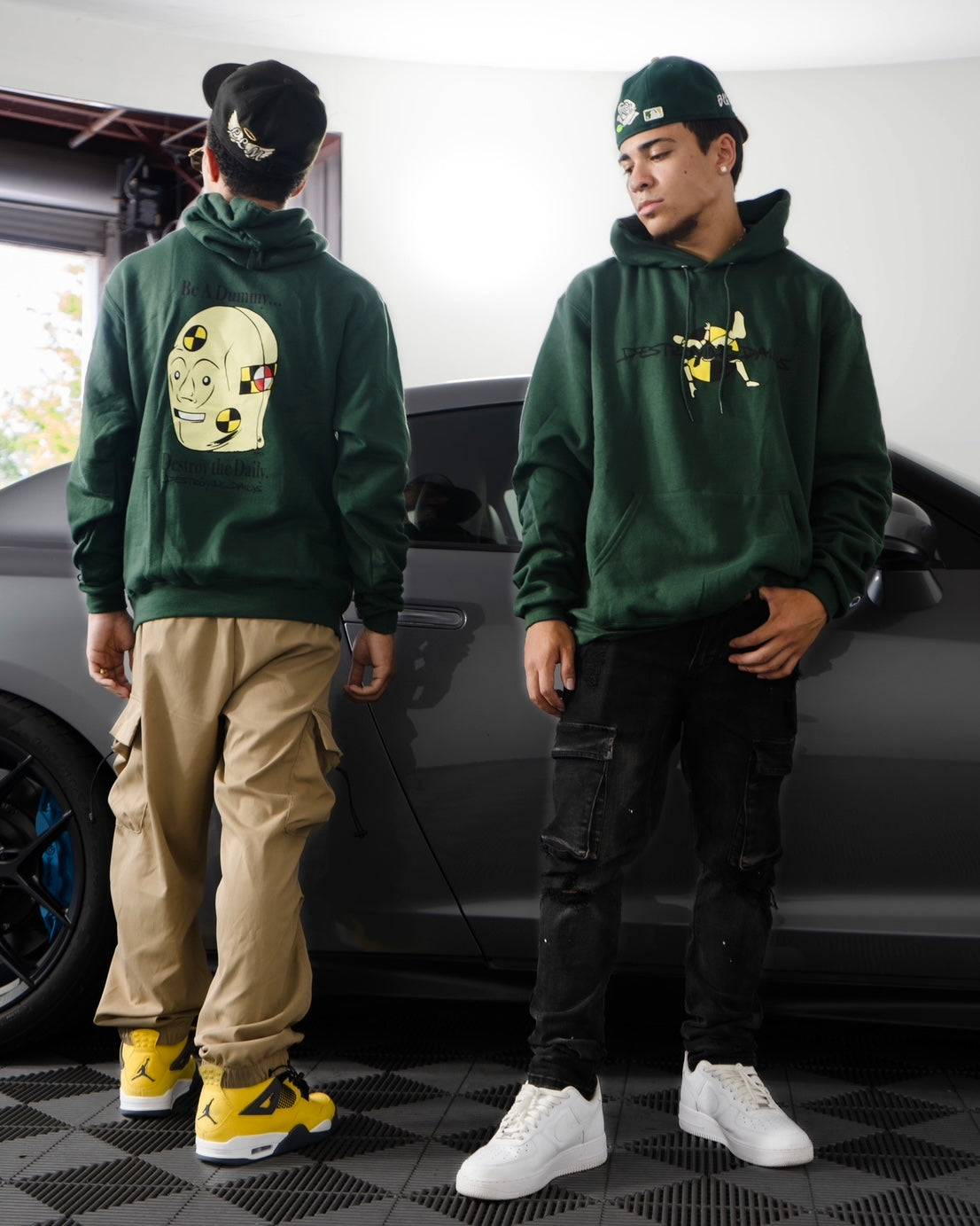 Male models with JDM Car wearing Dark green hoodie made of 50/50 cotton and polyester blend featuring a hand-drawn crash test dummy with the text "Be A Dummy... Destroy the Daily: Destroying Dailys," perfect for modified car enthusiasts.