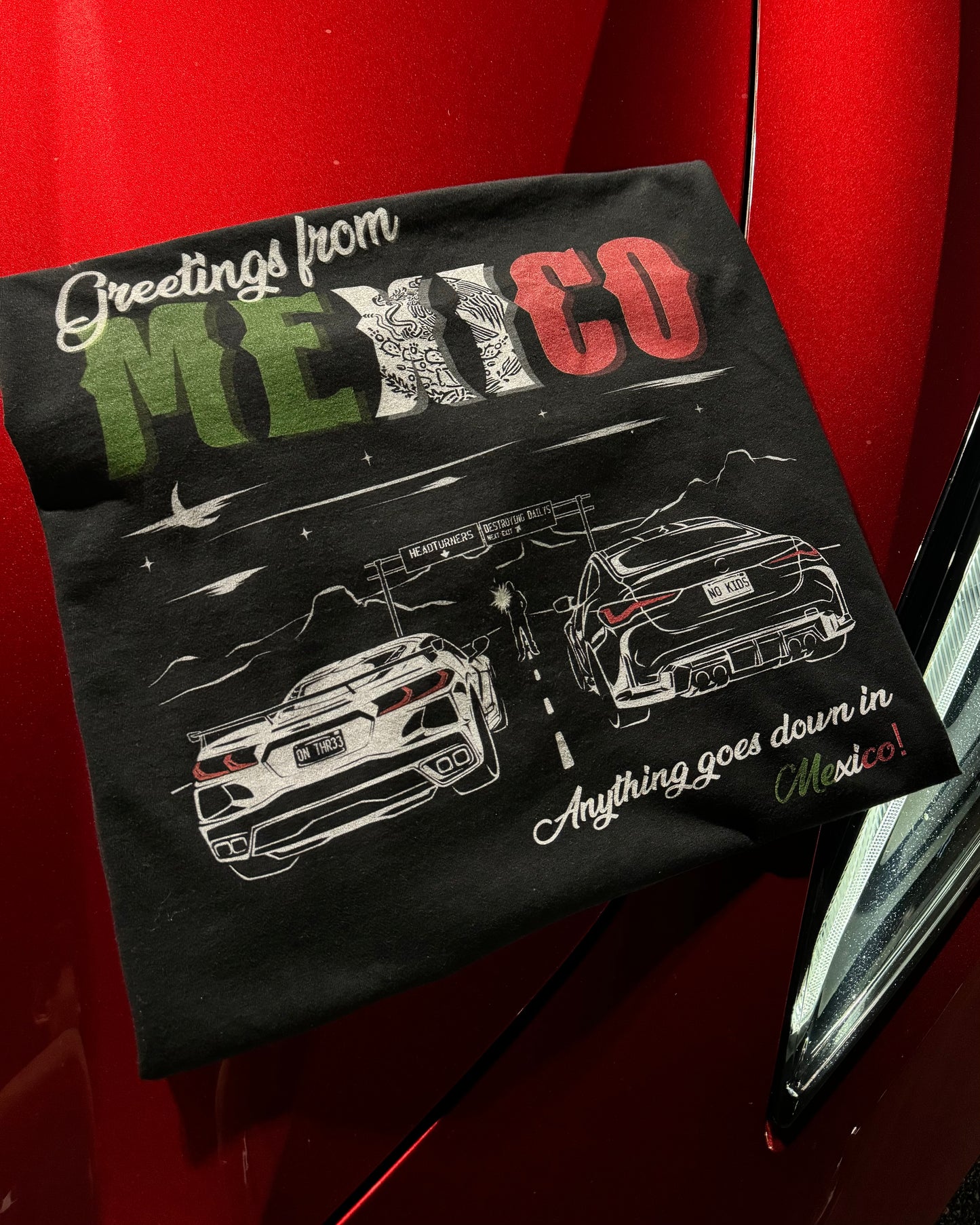 Black 100% cotton t-shirt featuring a hand-drawn Mexico racing scene with the text "Greetings from Mexico, Anything goes down in Mexico!" perfect for car enthusiasts. Mexico Racing T Shirt. Mexico racing corvette shirt. Mexico racing tee on a corvette