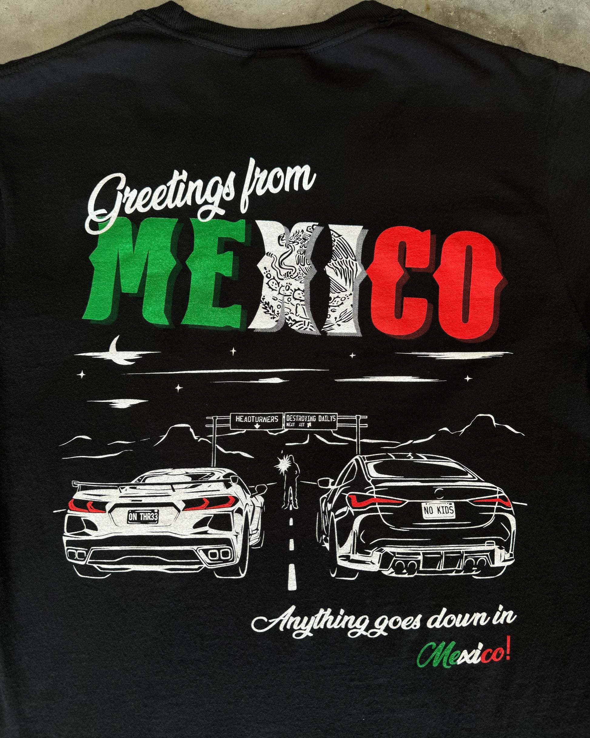 Black 100% cotton t-shirt featuring a hand-drawn Mexico racing scene with the text "Greetings from Mexico, Anything goes down in Mexico!" perfect for car enthusiasts. Mexico Racing T Shirt. Mexico racing corvette shirt. Detailed photo of back of tee